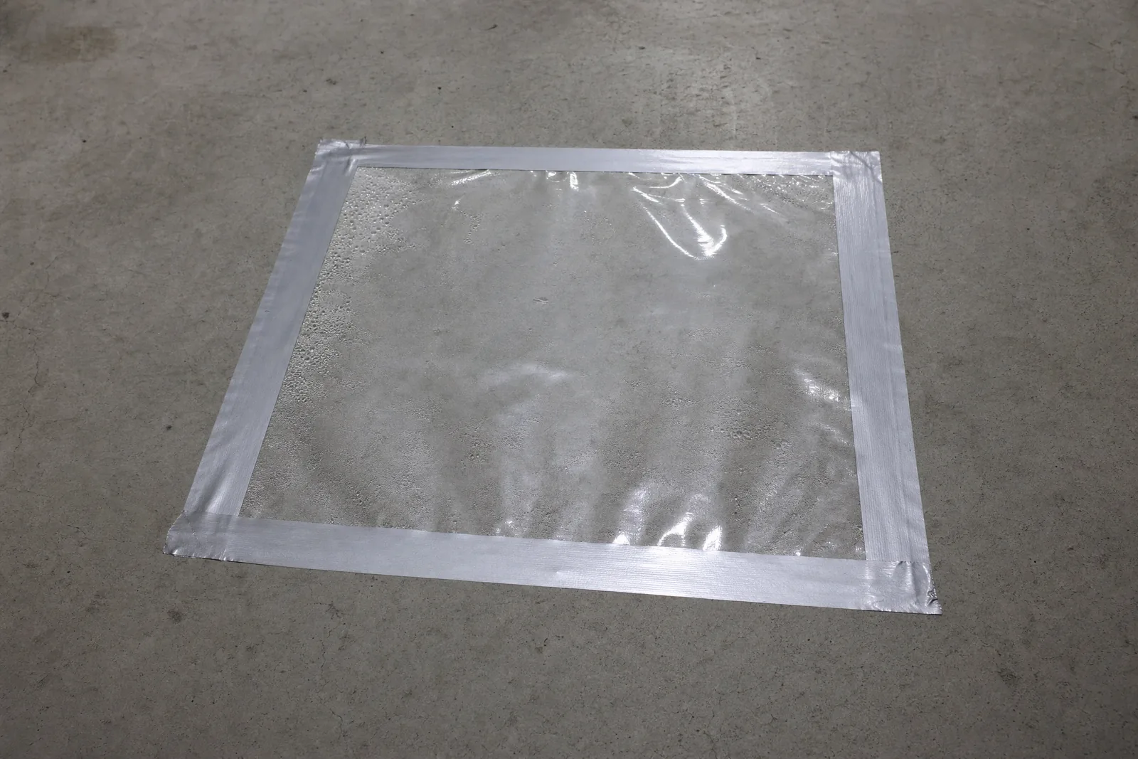 Clear plastic sheet taped flat to a bare concrete floor with condensation forming underneath